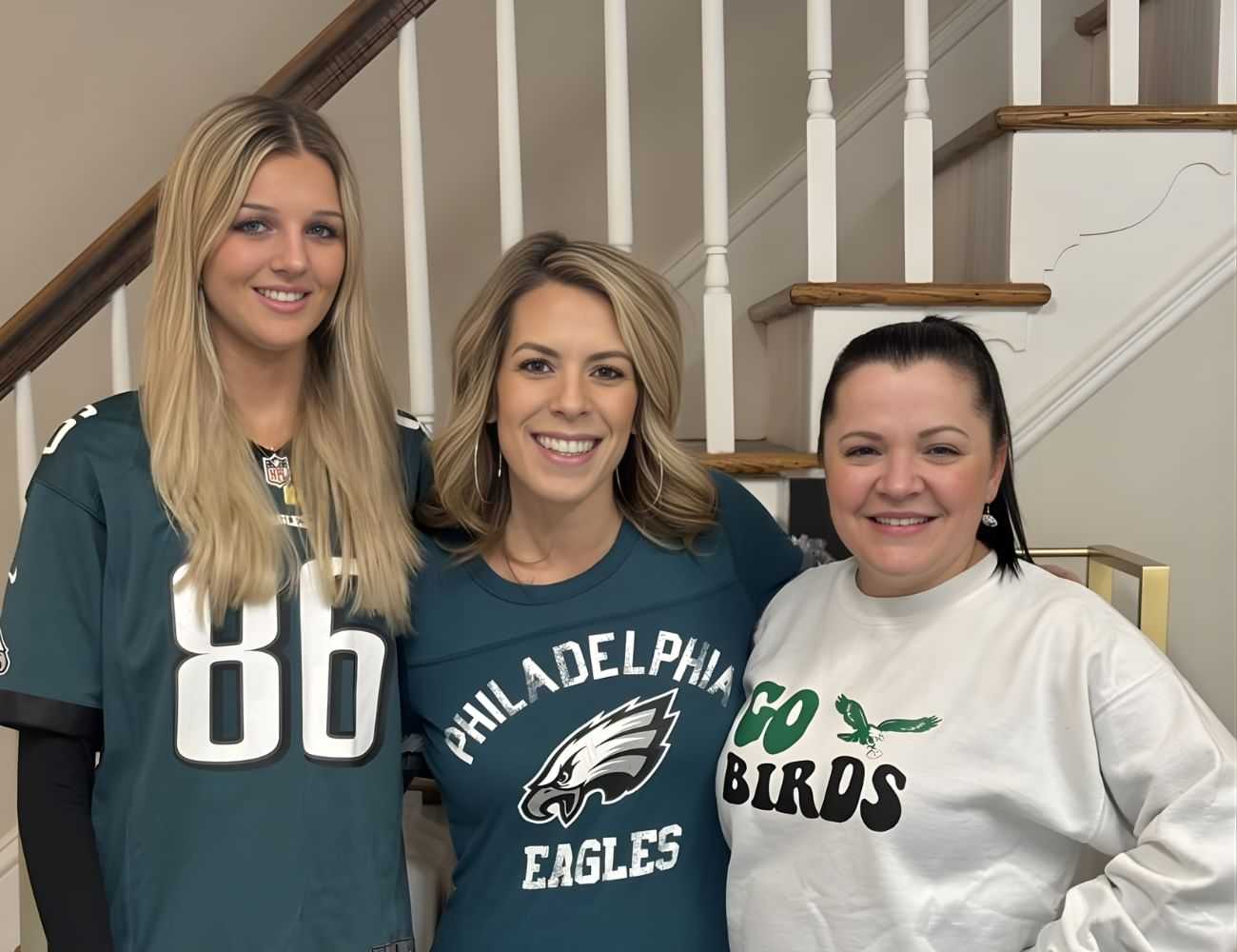 Three women in Eagles shirts smiling, standing by a staircase indoors.